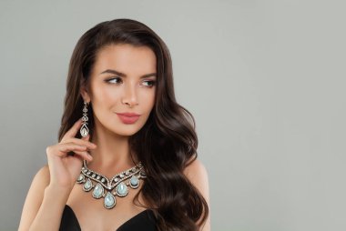 Uzun dalgalı saçlı güzel esmer kadın, gri arka planda elmas küpe ve kolye, moda güzelliği portresi.