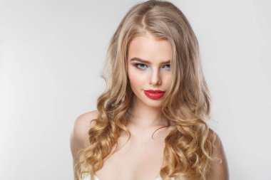 Makyajlı, uzun ve sağlıklı sarı dalgalı saçlı, beyaz stüdyo duvarına poz veren muhteşem bir kadın. Duygusal moda güzellik portresi