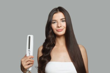 Çekici, genç bir kadın saç ütüsü kullanıyor ve uzun kahverengi saçlarını beyaz arka planda düzleştiriyor. Stüdyo moda güzellik portresi. Saç ütüleme ve saç stili konsepti