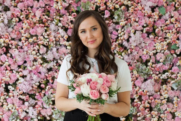 Young modest happy brunette woman with cute smile holding flower gift and posing on pink floral background.  Blooming, love, 8 march birthday holiday concept