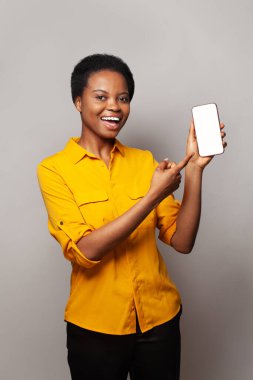 Akıllı telefonu boş ekran görüntüsüyle gösteren öğrenci bir kadın. Sarı gömlekli, gri stüdyo arka planında telefon olan bir model.