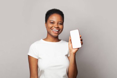 Akıllı telefonu boş ekran ekranıyla gösteren güzel bir kadın. Gri stüdyo arka planında telefon olan beyaz tişörtlü bir model.