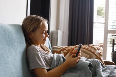 Çocuk, telefon uygulamaları kullanarak koltukta oturan cep telefonu cihazına bakıyor. Akıllı telefonu elinde tutan çocuk evde tek başına oyun oynuyor, çocuk ve cihaz kavramı
