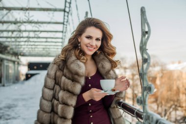 Elinde kahve tutan, kürk giyen, gülümseyen esmer kadın.