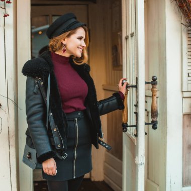 Siyah deri ceketli, siyah şapkalı esmer kadın dükkan kapısının yanında duruyor.