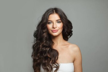Doğal güzellik. Çekici, neşeli, esmer, temiz tenli, güzel dalgalı saçlı ve doğal makyajlı bir kadın gri arka planda poz veren kameraya bakıyor.