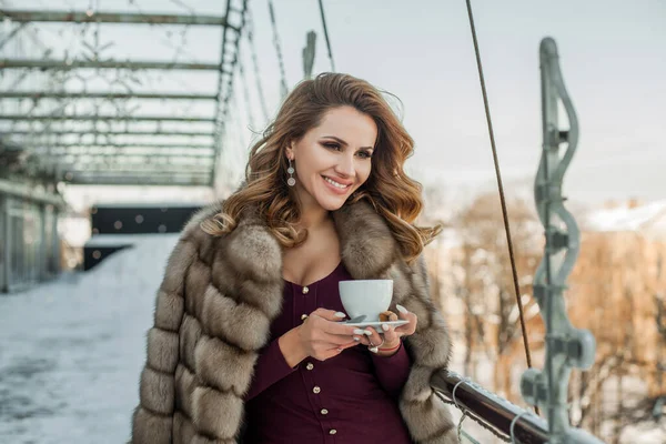 Kışlık kürk giyen güzel esmer kadın elinde bir fincan sıcak kahve tutuyor.