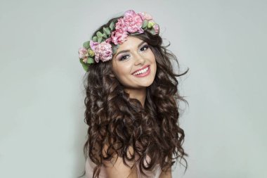 Makyajlı, uzun dalgalı saçlı, sağlıklı cilt ve beyaz arka planda çiçekler olan neşeli, genç esmer kadın.