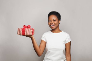 Beyaz tişörtlü, beyaz tişörtlü mutlu bir kadın beyaz stüdyo duvarına karşı sürpriz bir hediye kutusu tutuyor.