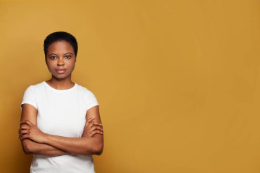 Kendine güvenen beyaz tişörtlü Afro-Amerikan kadın renkli sarı arka planda duruyor.