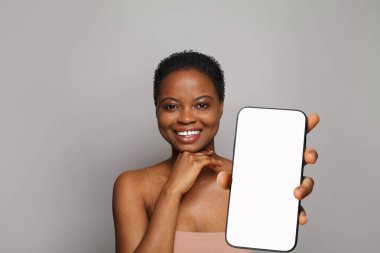 Neşeli, sağlıklı bir kadın gri stüdyo arka planında beyaz boş ekran ekranı olan akıllı telefonu tutuyor. Telefonlu mutlu model
