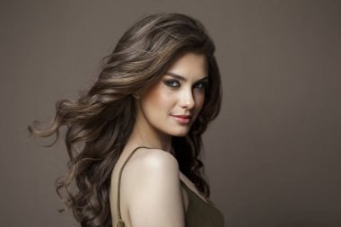 Uzun, sağlıklı dalgalı saçlı, sevimli gülüşlü ve makyajlı zarif bir kadın kahverengi stüdyo arka planında kameraya bakıyor, moda güzelliği portresi