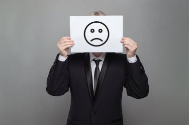 Anonim bir iş adamının portresi gri stüdyo arka planında üzgün bir ifade olan beyaz bir kağıdın arkasına saklanıyor.