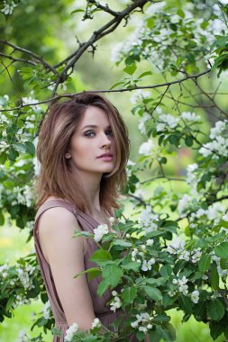 Taze ciltli, esmer, güzel, genç bir kadın model. Saç kesimi ve doğal makyajı, yeşil, bulanık ve bahar çiçekli yapraklara karşı.