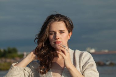 Neşeli esmer kadın taze tenli, doğal makyajlı ve sağlıklı dalgalı kahverengi saçlı güneşli sabahın ve deniz havasının tadını çıkarıyor. Kadın model açık hava moda portresi