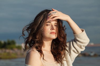 Dinlenen, kahverengi saçlı ve doğal makyajlı genç bir kadın model bir bahar sabahı tatil köyünün kıyısında yürüyor. Mutluluk, denge, sakinlik, duygu ve ruh hali konsepti. Gözler kapalı