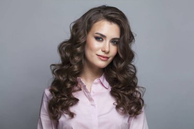 Taze tenli, uzun kıvırcık saçlı ve makyajlı güzel bir manken kadın gri arka planda poz veriyor.