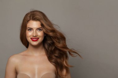 Mükemmel genç, mutlu, doğal makyajlı, temiz, sağlıklı ciltli ve gri arka planda uzun saçlı bir kadın modeli. Kıvırcık, saç bakımı, cilt bakımı ve yüz bakımı konsepti