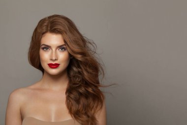 Doğal makyajlı, sağlıklı ve uzun saçlı, gri arka planda poz veren çekici, genç ve mutlu bir kadın modeli. Kıvırcık, saç bakımı, cilt bakımı ve yüz bakımı konsepti