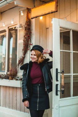 Siyah deri ceketli, siyah şapkalı esmer kadın dışarıda bekliyor.