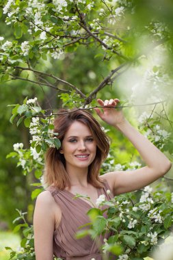 Genç, mutlu, esmer kadın model, taze cilt, saç kesimi ve doğal makyajla yeşil bulanık yapraklara karşı beyaz bahar çiçekli.