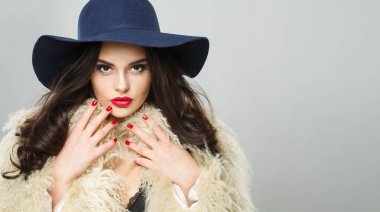 Parlak makyajlı, uzun dalgalı saçlı, mavi şapkalı, beyaz stüdyo duvarına poz veren esmer, yetişkin bir kadın model.