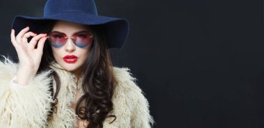 Parlak makyajlı, uzun dalgalı saçlı, mavi şapkalı ve güneş gözlüklü, mavi stüdyo arka planına poz veren genç yetişkin bir kadının stüdyo portresi.