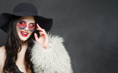Parlak makyajlı, uzun dalgalı saçlı, mavi şapkalı ve güneş gözlüklü, mavi stüdyo duvarına poz veren genç, yetişkin bir model.