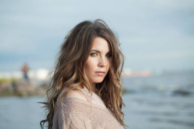 Mavi deniz sahili açıkhava portresi üzerinde doğal makyajlı ve uzun dalgalı kahverengi saçlı sevimli genç bir kadın.