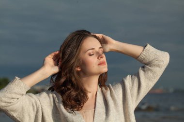 Kahverengi saçlı, doğal makyajlı, gülümseyen genç bir kadın model bir bahar sabahı tatil köyünün kıyılarında yürüyor. Mutluluk, denge, sükunet, duygu ve ruh hali konsepti