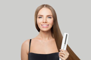 Güzel görünümlü esmer kadın düz saç ütüsünü tutuyor ve uzun parlak saçlarını beyaz arka planda düzleştiriyor. Saç bakım, saç stili, kuaförlük ve saç şekillendirme konsepti