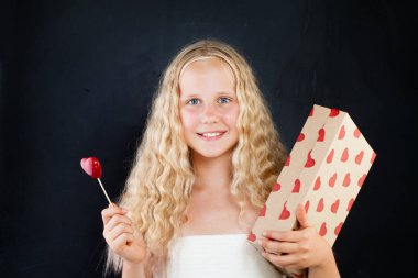 Gülümseyen genç kız elinde kırmızı kalple bir hediye kutusu tutuyor. Siyah arka planda uzun, sağlıklı kıvırcık saçlı sarışın çocuk.