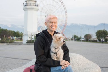 Hoş, orta yaşlı bir kadın, şehir parkında sevimli bir Jack Russell Terrier köpeği tutuyor. Açık hava portresi.