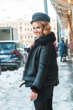 Kar şehrinin caddesinde duran siyah deri ceketli, mutlu gülümseyen esmer kadının portresi.