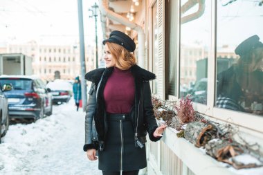 Siyah deri ceketli, siyah şapkalı, mavi çantalı esmer kadın kış şehrinin arka bahçesinde duruyor.