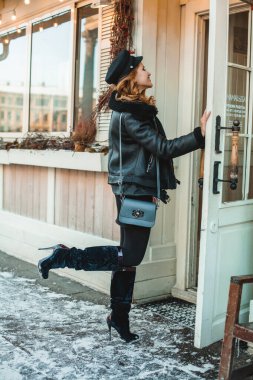 Siyah deri ceketli ve yüksek topuklu güzel esmer kadın. Kış kar şehrinin arka planında mavi çantalı..