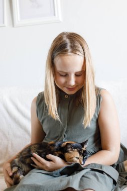 Gülümseyen genç kız kedi evcil hayvanını kucaklıyor rahat koltukta oturuyor, evde dinleniyor. Çocuk ve evcil kedi evde. Aşk, bağlılık ve ev hayatı kavramı.