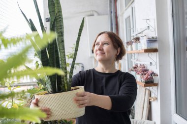 Büyüleyici orta yaşlı bir kadın elinde yeşil çiçekle evde dikiliyor. Bahçe işleri, rahatlama ve ekme konsepti.