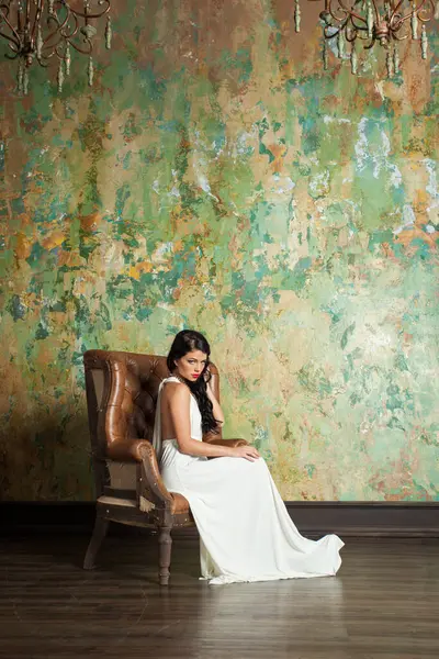 Uzun saçlı ve makyajlı görkemli bir kadın lüks bir iç mekanda oturuyor..