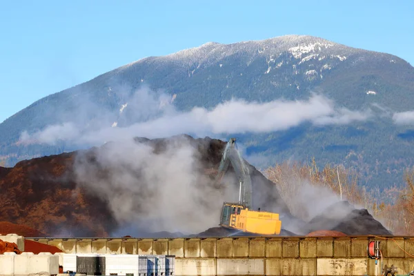 An industrial shovel moves a mountain of soil with a forested mountain in the background.