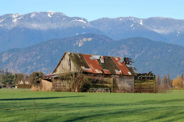 Full, profile view of the remnants of an old, turn-of-of-the-century farm building still standing in the valley as modern housing encroaches in the background.