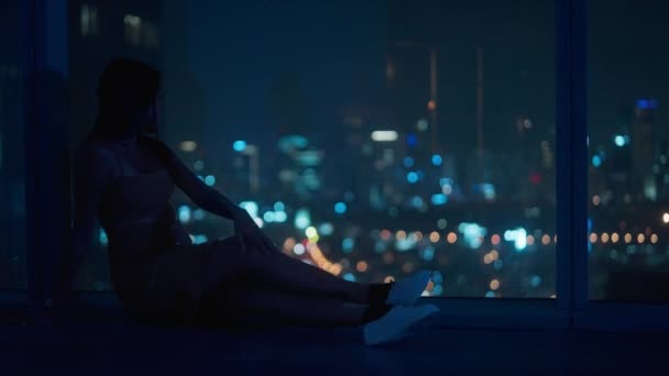 背の高い窓に座って夜の街の景色を楽しむ若い女性 ボケスローモーションショット — ストック動画