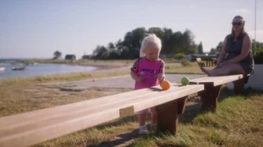 Annem pembe çiçekli küçük kızı izliyor. Arka planda manzara ve deniz olan ahşap bankta turuncu yuvarlanan topla oynuyor.