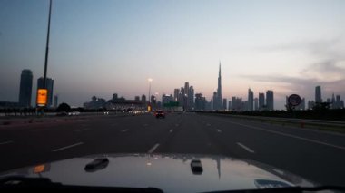 Dubai 'nin görkemli gökdelenlerini gören bir otoyolda gün batımında giden araba.