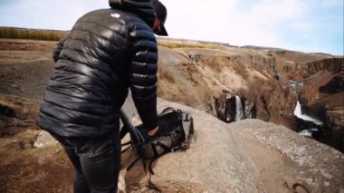 Ceket giyen ve kep takan bir adam çantasını açar. Güzel ufukta bir şelale manzaralı bir tepedeki büyük bir kayanın üzerine yerleştirilir..