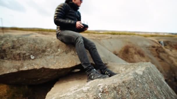 一个男人坐在岩石顶上 一边举着无人机 一边检查遥控器的倾斜画面 — 图库视频影像