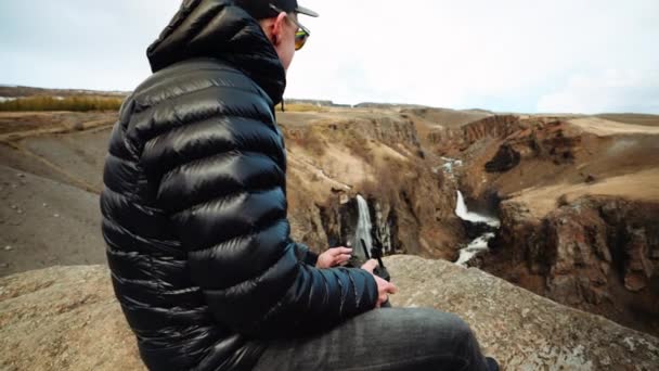 カメラがゆっくりと地平線の滝の明確なビューで彼の手のドローンのリモコンにズームとして岩の上に座ってジャケットを着て男の側面 — ストック動画
