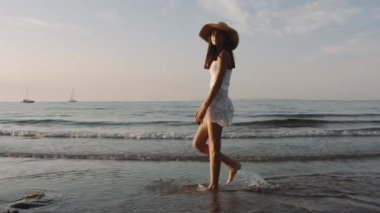 Hasır şapkalı genç bir kızın ufuk çizgisinde teknelerle sahil kenarında yürüdüğü geniş bir kare.