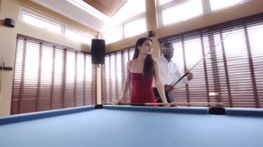 Bir adam bilardo masasına yaklaşıyor ve ıstakasıyla bilardo topunu hedef alıyor.