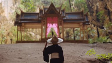 Kimono giymiş bir kadının bambu şapkasına dokunup Kuha Karuhas Pavyonu 'nun önünde dikilirken çekilen görüntüsü.